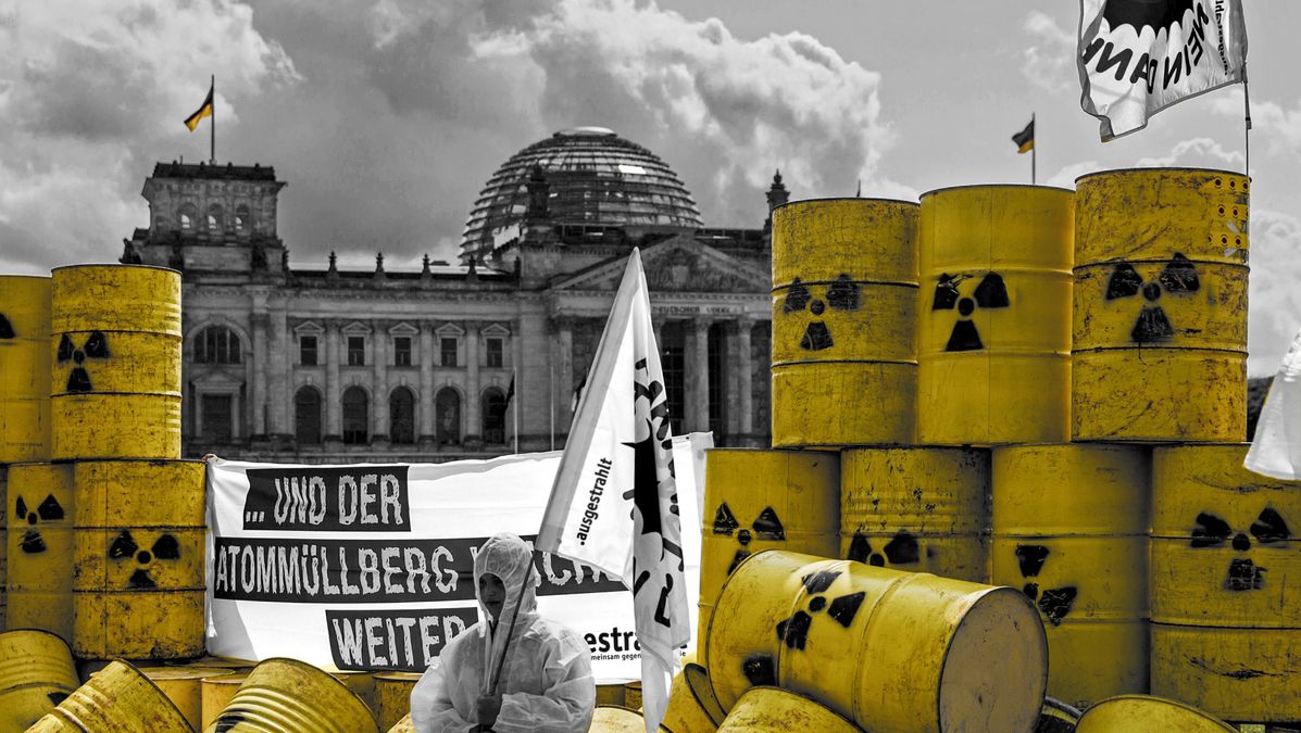 Gelbe Fässer vor dem Bundestag und ein Mensch mit einer Fahne und hinter ihm hängt ein Transparent mit Aufschrift.